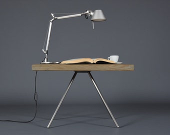 May include: A silver desk lamp with a flexible arm is on a wooden table with a book and a white cup. The table has three silver legs.