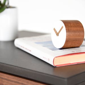 May include: A white and brown wooden clock with a simple design. The clock is sitting on a book with a red spine.