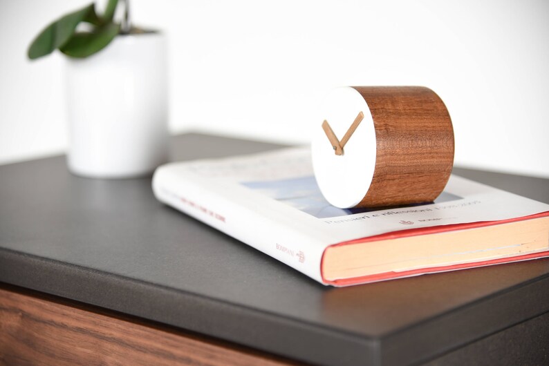 May include: A white and brown wooden clock with a simple design. The clock is sitting on a book with a red spine.