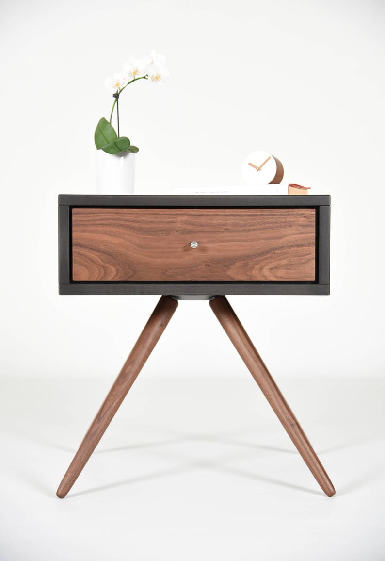 May include: A dark gray nightstand with a single drawer made of walnut wood. The nightstand has three wooden legs and a white orchid plant on top.
