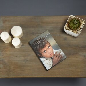 May include: A wooden table top with three white candles, a book titled "IL BAMBINO CHE CREDEVA NELLA LIBERTA" by Bina Shah, and a small white planter with a green cactus.
