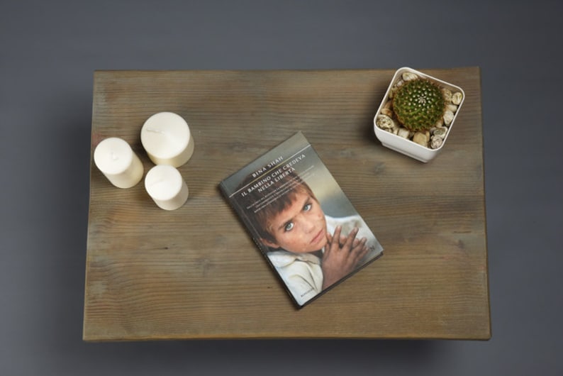 May include: A wooden table top with three white candles, a book titled "IL BAMBINO CHE CREDEVA NELLA LIBERTA" by Bina Shah, and a small white planter with a green cactus.