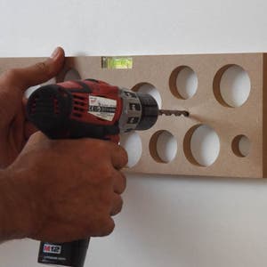 May include: A person is using a red and black power drill to drill holes in a piece of wood with a level attached to the top.