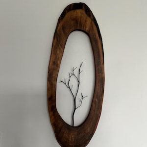 Może przedstawiać: Rustykalne, owalne drewniane dzieło sztuki na ścianę. Rama jest w bogatym brązowym kolorze z naturalnym usłojeniem drewna. Wewnątrz ramy znajduje się czarna metalowa sylwetka drzewa, tworząca element dekoracyjny inspirowany naturą.