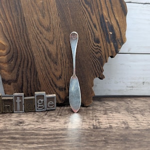 Può includere: Un coltello da burro d'argento con un manico decorativo è appoggiato su una superficie di legno. La parola "vintage" è scritta con blocchi di lettere individuali. Un tagliere di legno a forma di stato è sullo sfondo.