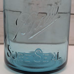 May include: A vintage blue glass jar with the embossed word "Ball" and "Sure Seal". The jar has a classic design, with a wide mouth and a slightly tapered body. The glass has a slight blue tint, and the embossed lettering is clear and easy to read.