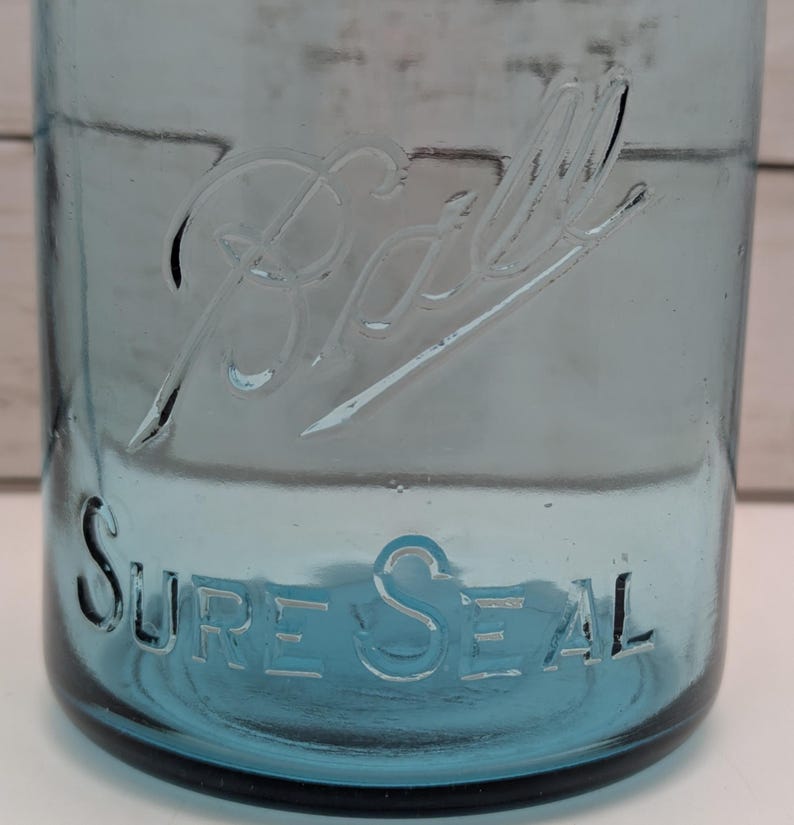 May include: A vintage blue glass jar with the embossed word "Ball" and "Sure Seal". The jar has a classic design, with a wide mouth and a slightly tapered body. The glass has a slight blue tint, and the embossed lettering is clear and easy to read.