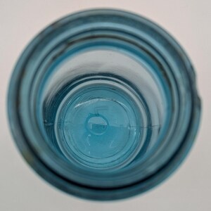 May include: An overhead view of a clear, blue glass bottle. The bottle has a wide mouth and a rounded base. The glass has a slightly textured surface. The bottle is empty and sits on a white surface.