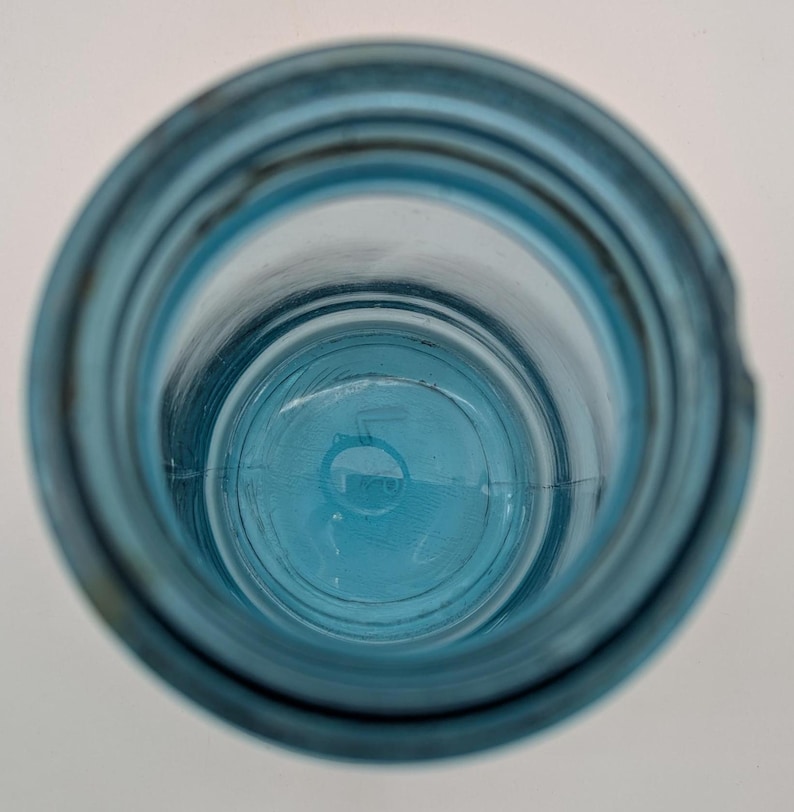 May include: An overhead view of a clear, blue glass bottle. The bottle has a wide mouth and a rounded base. The glass has a slightly textured surface. The bottle is empty and sits on a white surface.