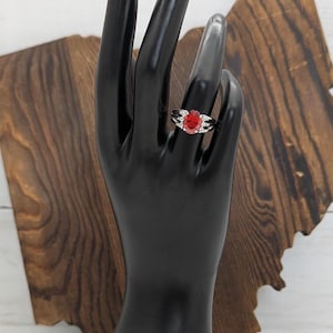 May include: A silver-toned ring featuring a prominent oval-cut red gemstone at the center, flanked by small clear stones on each side. The band has a split design, and the ring is displayed on a black mannequin hand model.