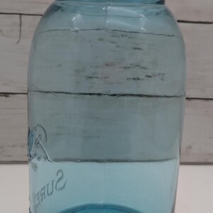 May include: A vintage blue glass jar with a matching lid. The jar has a wide mouth and a rounded body. The word "Sure" is embossed on the side. The jar is likely used for canning or storage.