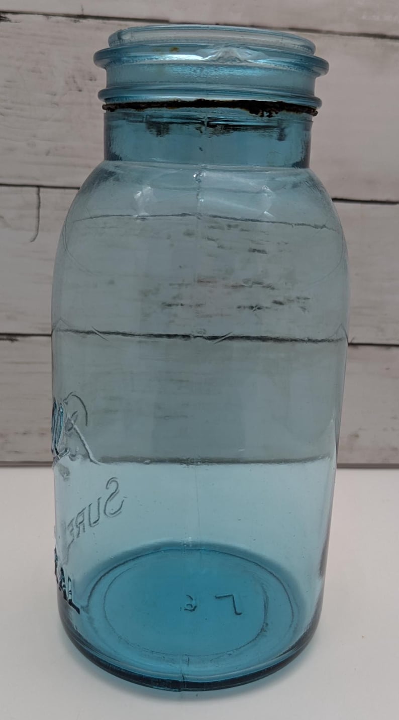 May include: A vintage blue glass jar with a matching lid. The jar has a wide mouth and a rounded body. The word "Sure" is embossed on the side. The jar is likely used for canning or storage.