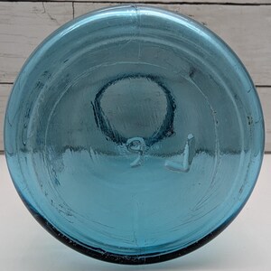 May include: A clear, blue glass bottle with a rounded base. The bottom of the bottle has a circular indentation and the number "9" and the letter "L" are embossed on the base. The bottle is made of thick glass.