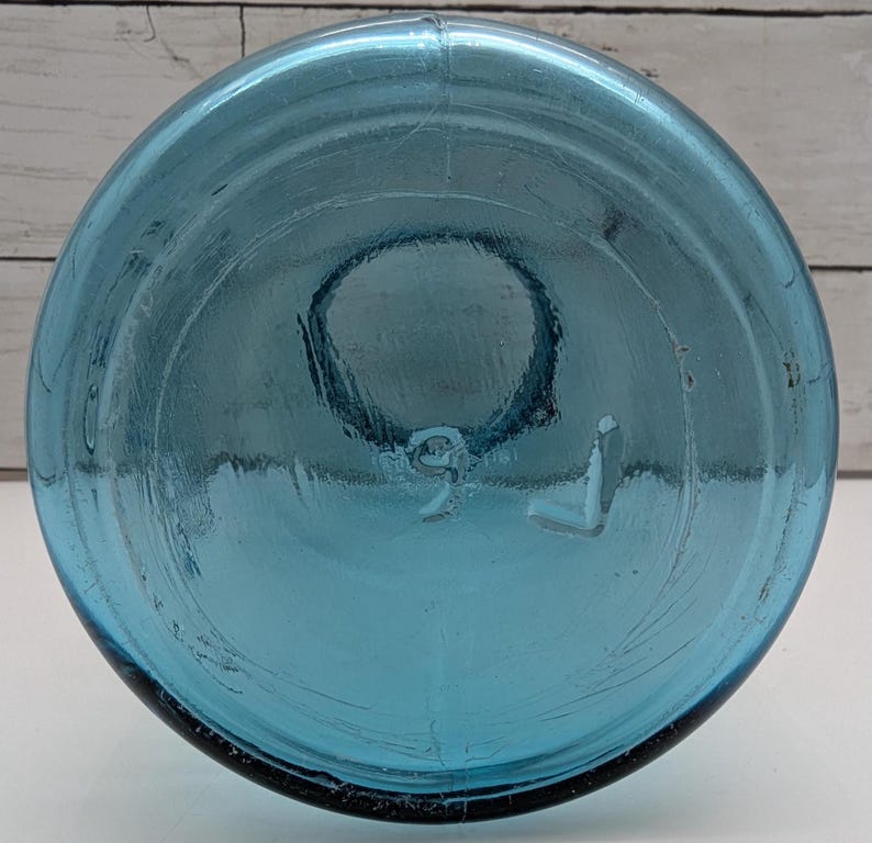 May include: A clear, blue glass bottle with a rounded base. The bottom of the bottle has a circular indentation and the number "9" and the letter "L" are embossed on the base. The bottle is made of thick glass.