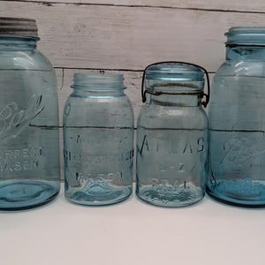 May include: A collection of vintage blue glass Ball and Atlas canning jars with metal lids. The jars vary in size and shape, with embossed lettering and a classic design. The jars are arranged on a white surface against a wooden background.