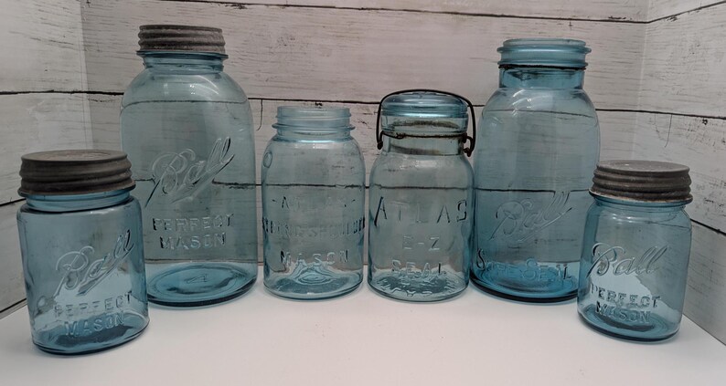 May include: A collection of vintage blue glass Ball and Atlas canning jars with metal lids. The jars vary in size and shape, with embossed lettering and a classic design. The jars are arranged on a white surface against a wooden background.