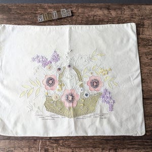 以下が含まれることがあります： クリーム色の長方形枕カバー。刺繍された花柄のデザイン。ピンクと紫の花でいっぱいのバスケット、緑の葉と緑のバスケットが特徴です。「vintage」の文字が上部に刻印されています。