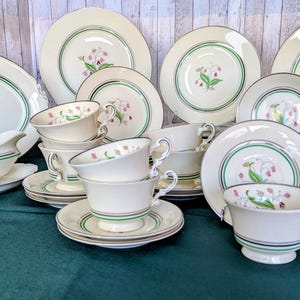 May include: A collection of vintage white porcelain dinnerware with a floral design. The set includes plates, cups, saucers, a serving platter, and a gravy boat. Each piece features a green and gold trim and a pink and green floral motif.