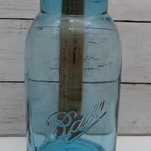 May include: A large, blue glass Ball jar with a wide mouth and a screw-on lid. The jar has the Ball logo embossed on the front. A wooden ruler is inside the jar, measuring approximately 10 inches.