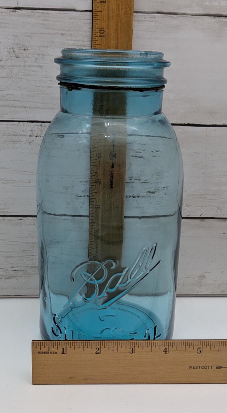 May include: A large, blue glass Ball jar with a wide mouth and a screw-on lid. The jar has the Ball logo embossed on the front. A wooden ruler is inside the jar, measuring approximately 10 inches.