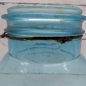 May include: Close-up of a vintage, light blue glass jar with a matching lid. The jar has a wide mouth and a metal ring around the neck. The lid and ring show signs of age and wear, with some rust visible.
