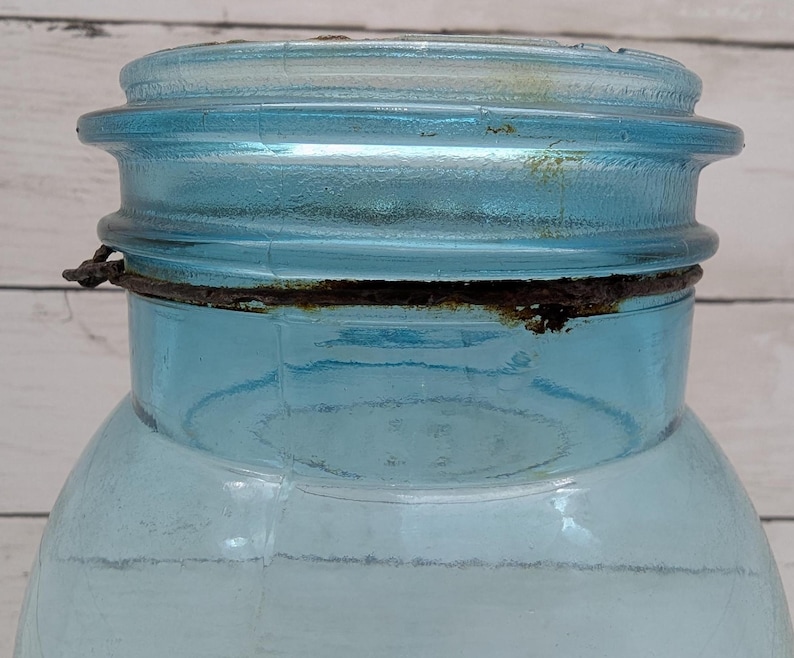 May include: Close-up of a vintage, light blue glass jar with a matching lid. The jar has a wide mouth and a metal ring around the neck. The lid and ring show signs of age and wear, with some rust visible.