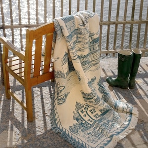 May include: A wooden chair with a blue and white throw blanket draped over it. The blanket features a lighthouse and other nautical imagery. The chair is on a stone patio overlooking a body of water.