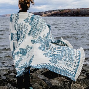 May include: A white and blue woven throw blanket with a nautical design featuring a lighthouse and buildings. The blanket has a rope trim and fringe edges.
