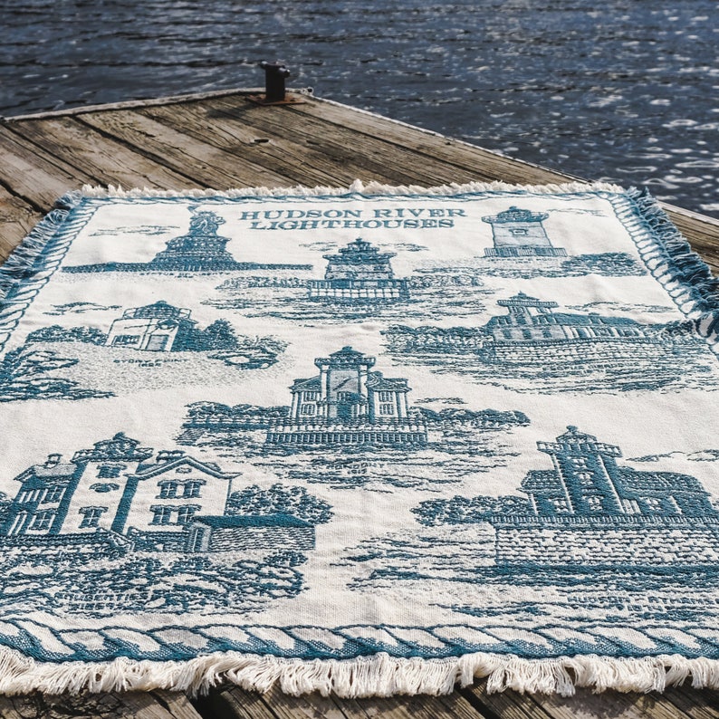 May include: A white woven throw blanket with a blue design featuring lighthouses along the Hudson River. The text "HUDSON RIVER LIGHTHOUSES" is printed in blue at the top of the blanket.