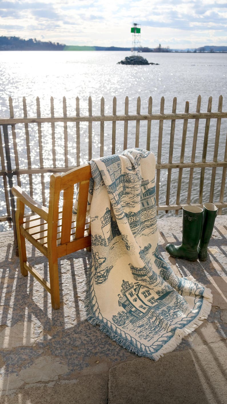 May include: A wooden chair with a blue and white throw blanket draped over it. The blanket features a lighthouse and other nautical imagery. The chair is on a stone patio overlooking a body of water.