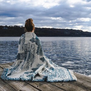 May include: A person sitting on a wooden dock, wrapped in a white and blue patterned blanket, looking out at a body of water. The sky is cloudy and the water is calm.