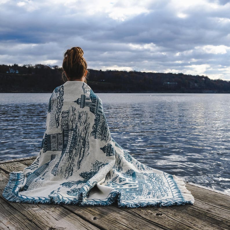 May include: A person sitting on a wooden dock, wrapped in a white and blue patterned blanket, looking out at a body of water. The sky is cloudy and the water is calm.