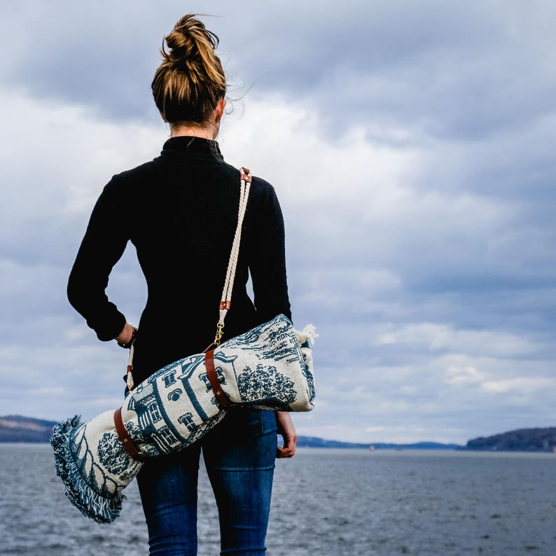 May include: A woman wearing a black turtleneck sweater and blue jeans is carrying a rolled up blue and white patterned blanket with a brown leather strap.