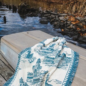May include: A white and blue throw blanket with a nautical theme. The blanket features a lighthouse, houses, and a rope border. The blanket is folded and lying on a wooden surface.