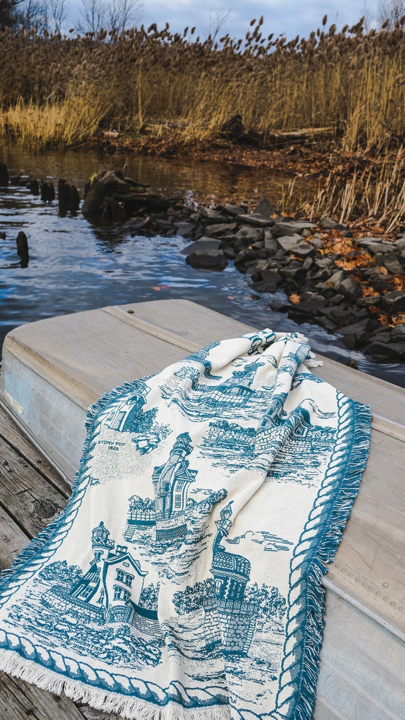 May include: A white and blue throw blanket with a nautical theme. The blanket features a lighthouse, houses, and a rope border. The blanket is folded and lying on a wooden surface.