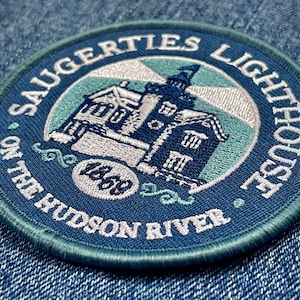 May include: A blue and white embroidered patch with the text "Saugerties Lighthouse on the Hudson River" and the year "1868" in a circle. The patch features a white and blue lighthouse with a white roof.