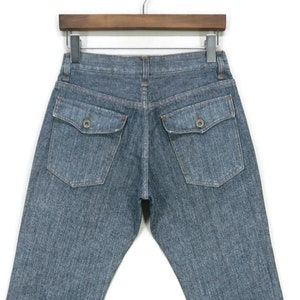 Puede incluir: Un par de pantalones vaqueros azules de mezclilla con dos bolsillos traseros. Los pantalones están colgados en una percha de madera.