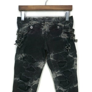Op de afbeelding: Zwarte denim jeans met een distressed, tie-dye effect. De jeans heeft meerdere scheuren en gaten, samen met metalen hardware en gespen aan de zijkanten. De jeans hangt aan een houten hanger tegen een witte achtergrond.
