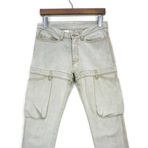 May include: Light gray denim pants with unique pocket detailing. The pants feature a distinctive design with additional pockets and seams, hanging from a hanger against a white background. The stitching is a contrasting tan color.