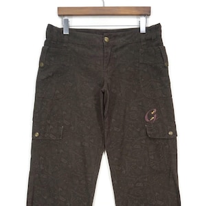 May include: Brown cargo pants with a paisley pattern and multiple pockets. The pants have a button and zipper closure, and a small embroidered "G" logo on one pocket. The pants are hanging on a wooden hanger.