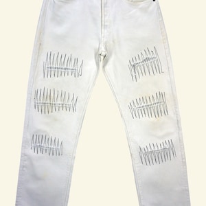 Pode incluir: Jeans brancos de ganga com detalhes costurados a preto. Os jeans apresentam um design clássico de cinco bolsos e são embelezados com filas de costuras pretas nos joelhos e coxas. Os jeans estão pendurados num cabide de metal.