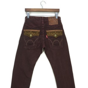 May include: A pair of brown denim jeans with embroidered details on the back pockets. The jeans have a brown leather patch with the text "EDWIN" on the back waistband.