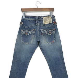 May include: A pair of blue denim jeans with red stitching detail. The jeans have a faded look and a label that reads "Blue Trip 503".