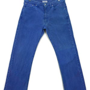 Puede incluir: Un par de vaqueros azules colgados de una percha de madera. Los vaqueros son de color azul medio liso con un diseño clásico de cinco bolsillos. Los vaqueros están hechos de un material de mezclilla duradero y tienen un corte de pierna recta.