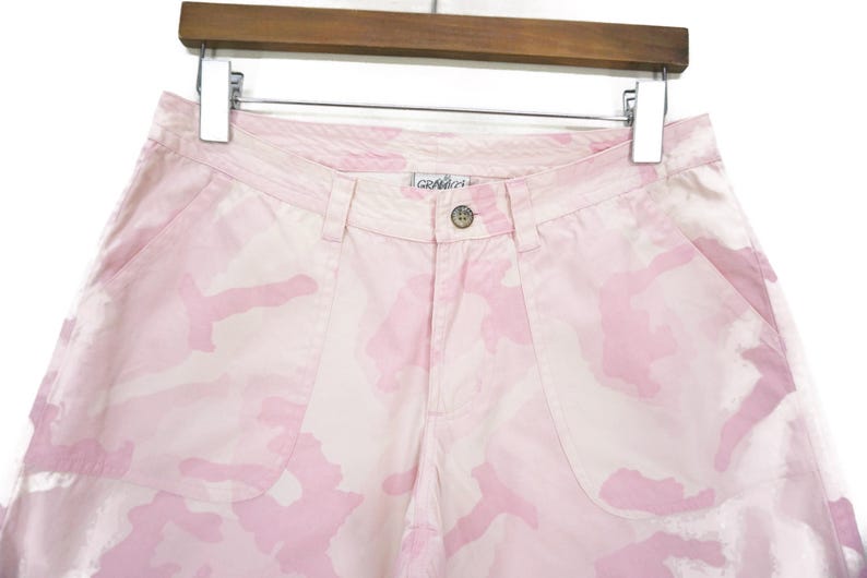 May include: Pink camouflage cargo shorts with a light pink color and a subtle camouflage pattern. The shorts have a button and zipper closure and belt loops.
