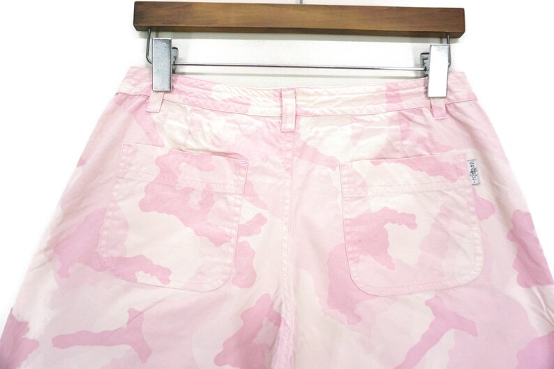 May include: Pink camouflage print shorts with two pockets. The shorts have a button and zipper closure.