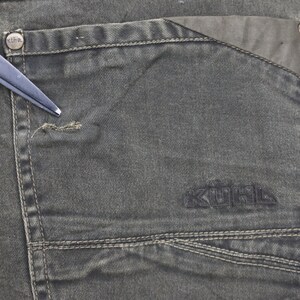 Puede incluir: Primer plano de un par de pantalones vaqueros de color verde oscuro con un bolsillo grande en el lado derecho. El bolsillo tiene una solapa y est&aacute; cosido con un hilo de contraste. La palabra "KOHL'S" est&aacute; bordada en negro en el bolsillo.