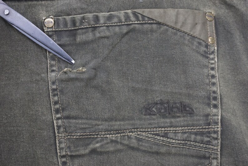 Puede incluir: Primer plano de un par de pantalones vaqueros de color verde oscuro con un bolsillo grande en el lado derecho. El bolsillo tiene una solapa y est&aacute; cosido con un hilo de contraste. La palabra "KOHL'S" est&aacute; bordada en negro en el bolsillo.