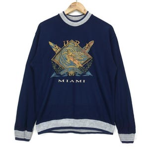 May include: Navy blue sweatshirt with a gray collar, cuffs, and hem. The front features a graphic of a surfer riding a wave, with the word "SURF" above and "MIAMI" below. The design includes crossed surfboards.