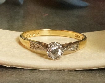 May include: A gold band engagement ring with a single round diamond set in a white metal setting. The band is decorated with small diamonds on each side of the center stone.